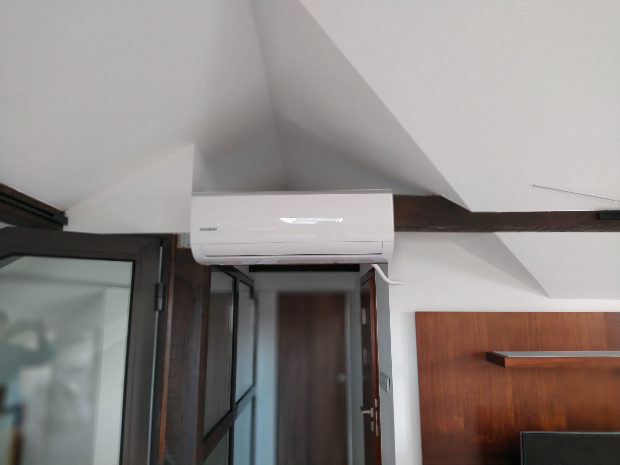 Biała jednostka wewnętrzna klimatyzacji marki Kaisai zamontowana pod skośnym sufitem, widoczny fragment drewnianej belki stropowej i drzwi balkonowe.