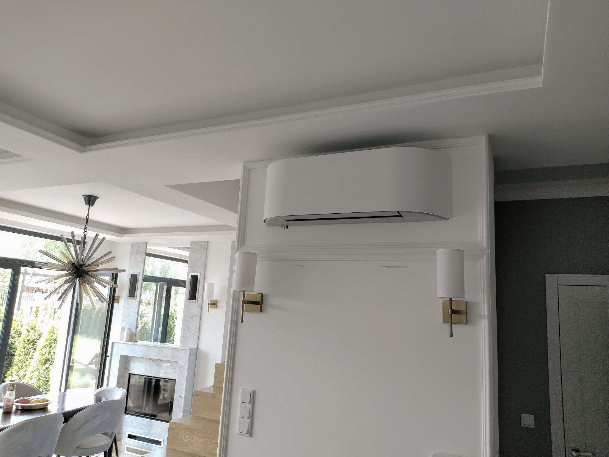 Nowoczesny klimatyzator ścienny w jasnym wnętrzu z kominkiem i widokiem na zieleń za oknem.