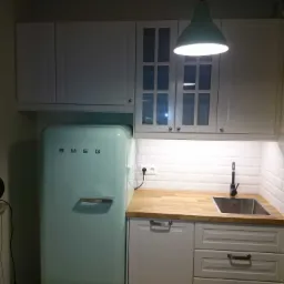 Kuchnia z białymi szafkami, drewnianym blatem i turkusową lodówką SMEG, oświetlona wiszącą lampą w kolorze miętowym.