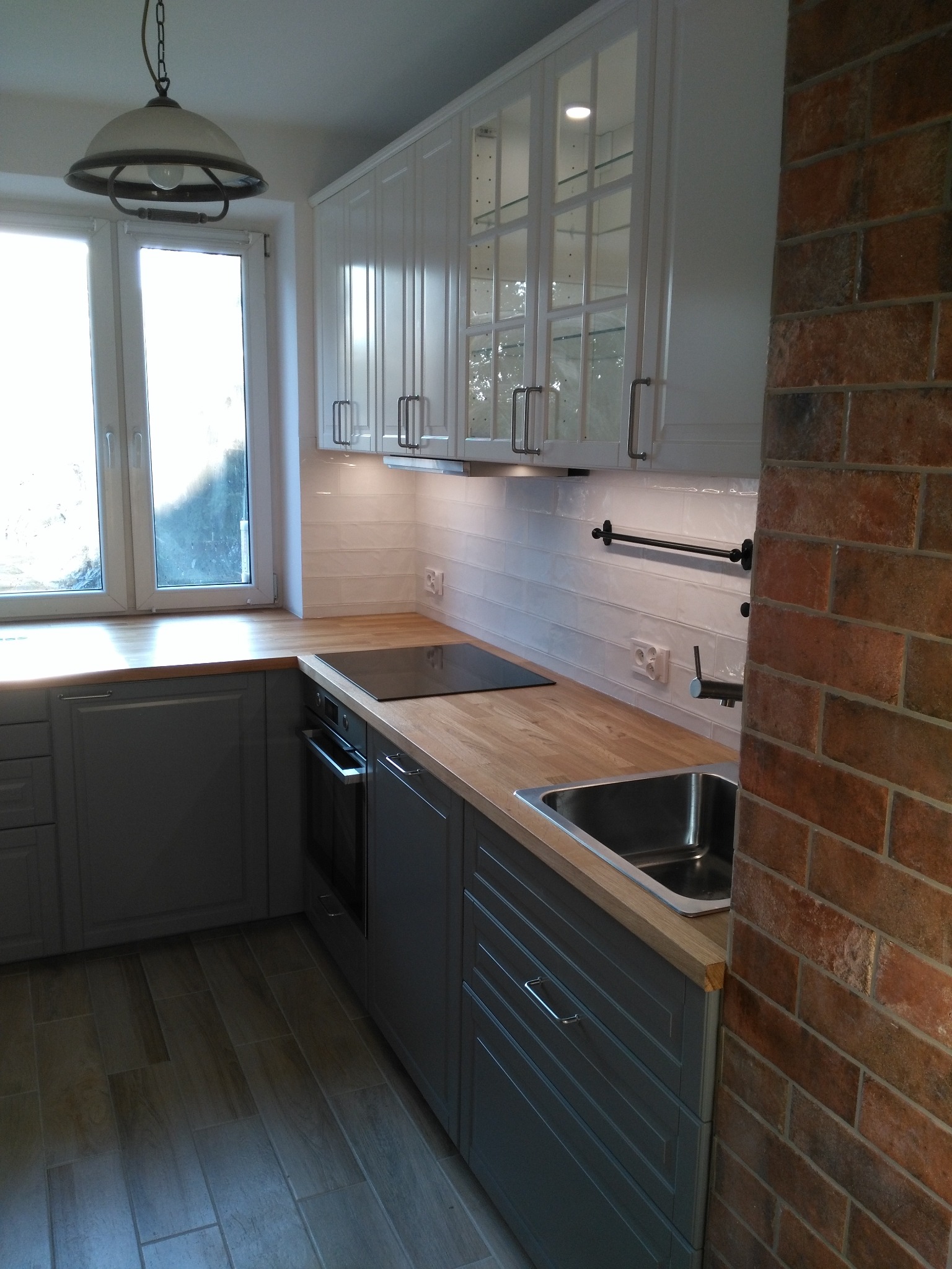 Szara zabudowa kuchenna z drewnianym blatem, białymi szafkami wiszącymi z przeszklonymi frontami, widok na okno i lampę wiszącą, fragment ceglanej ściany z prawej strony.