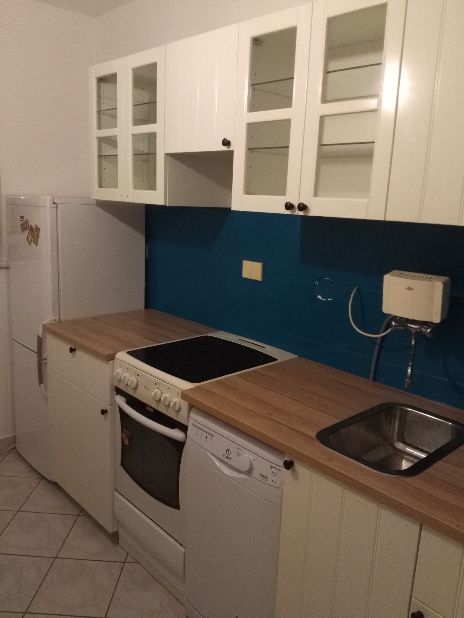 Biała kuchnia z drewnianym blatem i niebieskimi płytkami nad blatem, widoczny zlew, płyta grzewcza, piekarnik, zmywarka i lodówka.