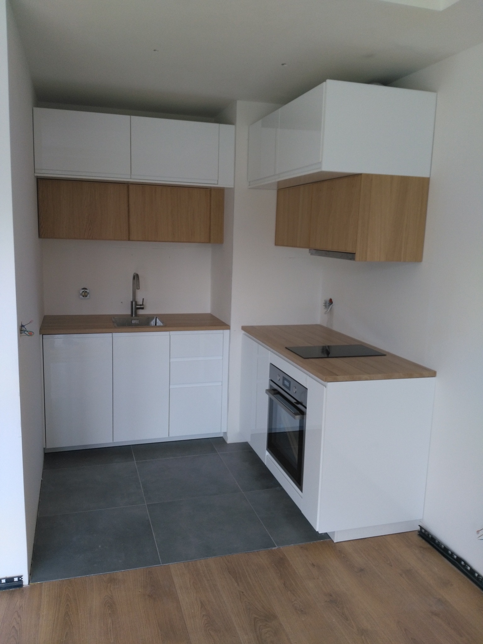 Nowoczesna, minimalistyczna kuchnia z białymi szafkami i drewnianymi blatami, z widocznym zlewozmywakiem, płytą indukcyjną i piekarnikiem, na podłodze szare płytki przechodzące w panele drewnopodobne.