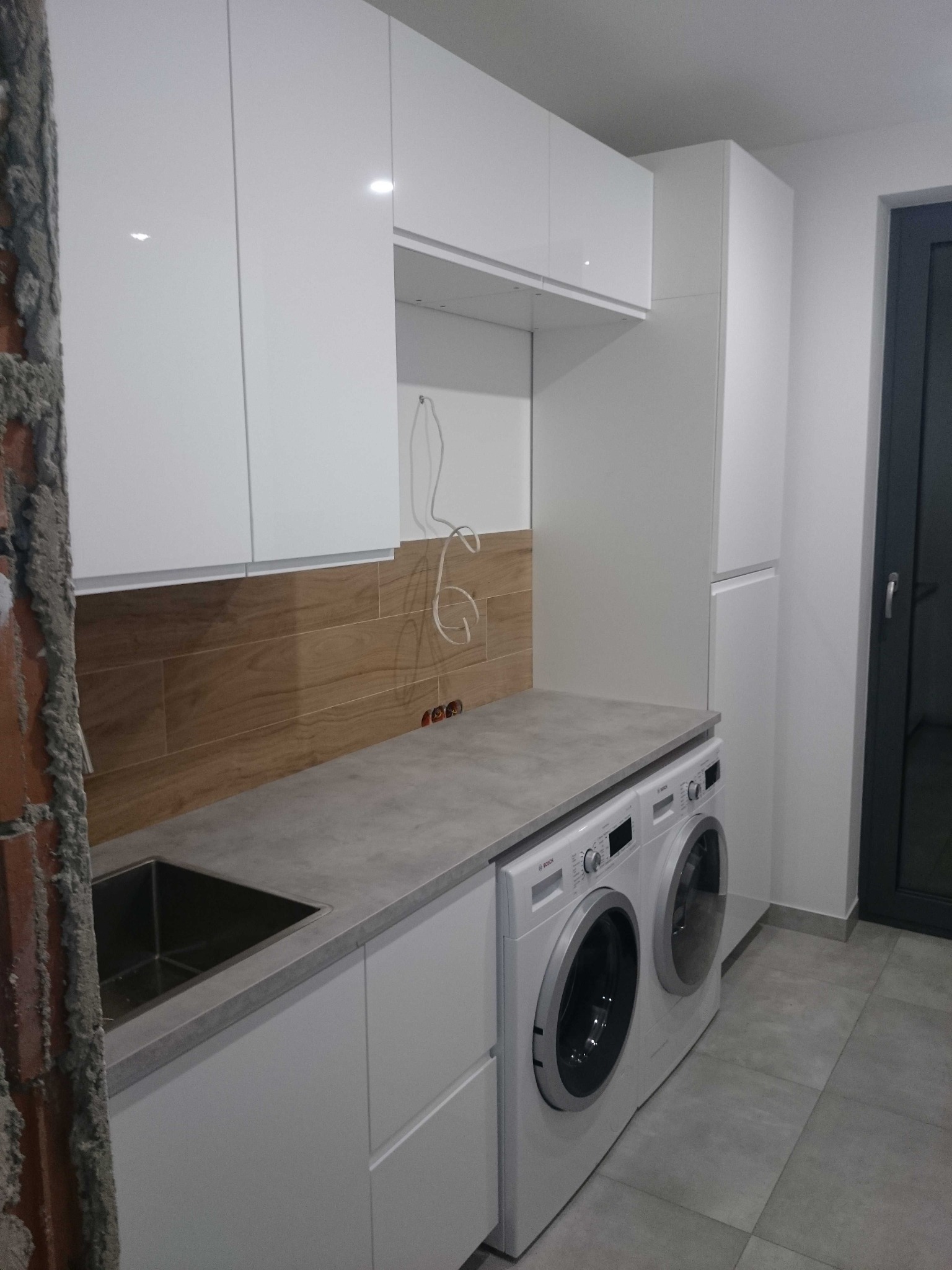 Białe szafki kuchenne z szarym blatem i drewnopodobnymi panelami ściennymi, z pralkami w zabudowie, widok na niedokończoną ścianę z cegły po lewej stronie.