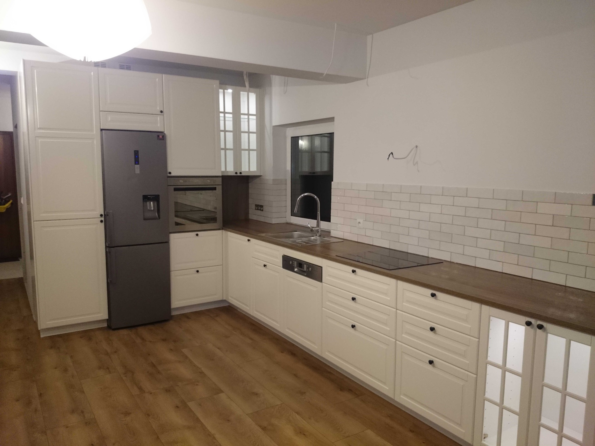 Wnętrze kuchni w trakcie montażu, widoczne białe szafki z ciemnymi gałkami, drewniany blat, białe płytki na ścianie i sprzęt AGD, przewody wystające z sufitu.