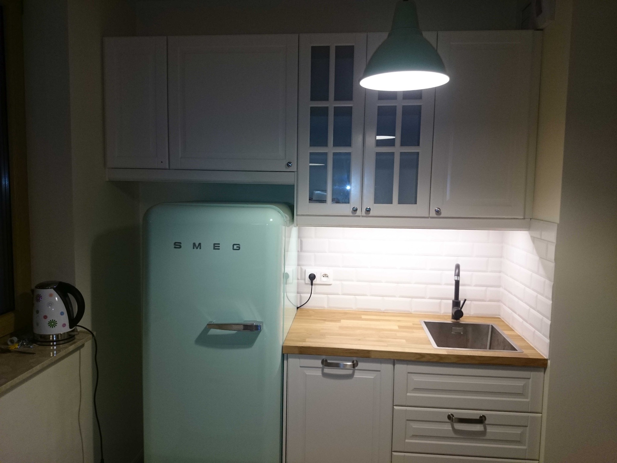 Kuchnia z białymi szafkami, drewnianym blatem i turkusową lodówką SMEG, oświetlona wiszącą lampą w kolorze miętowym.