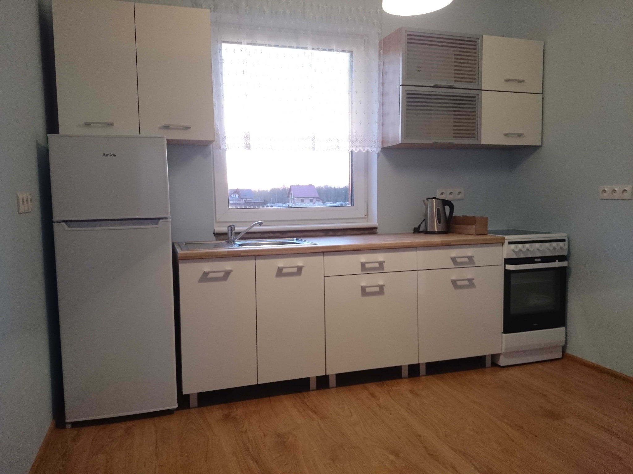 Jasna kuchnia z białymi frontami, widoczny zlew, płyta grzewcza i lodówka Amica, okno z widokiem na domy jednorodzinne.