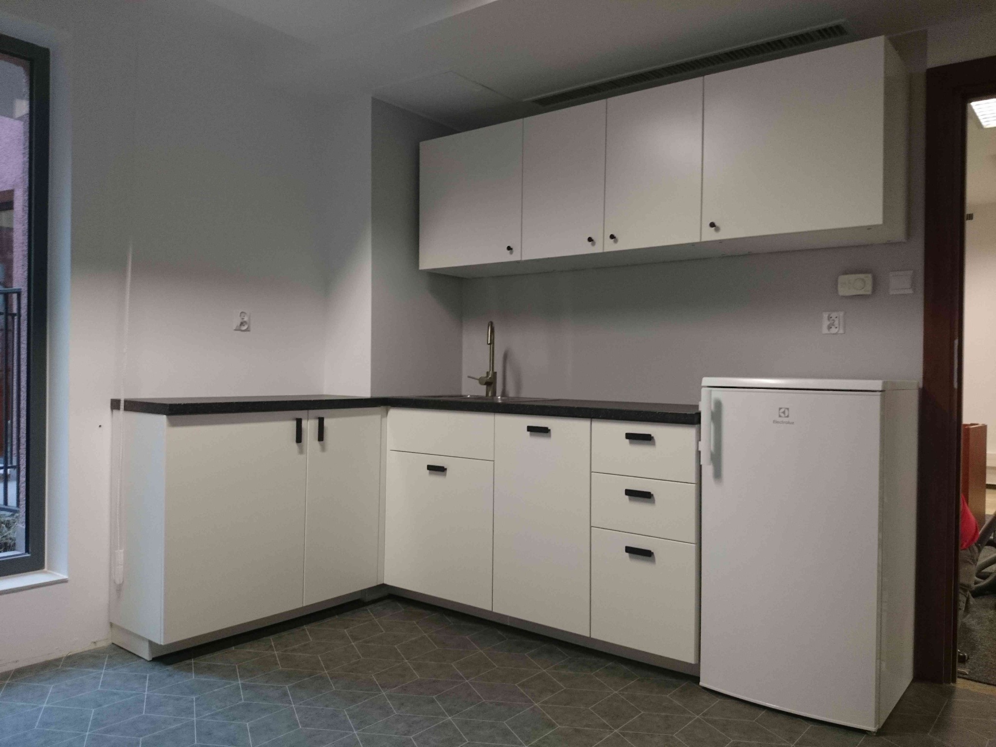 Wnętrze kuchni z białymi szafkami, czarnym blatem i białą lodówką Electrolux, widok na geometryczną podłogę i okno z boku.