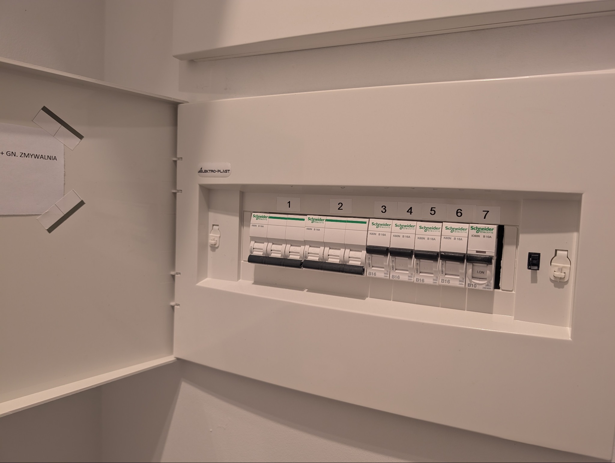 Otwarta, biała skrzynka elektryczna z siedmioma bezpiecznikami Schneider Electric, oznaczonymi numerami 1-7, w minimalistycznym wnętrzu.