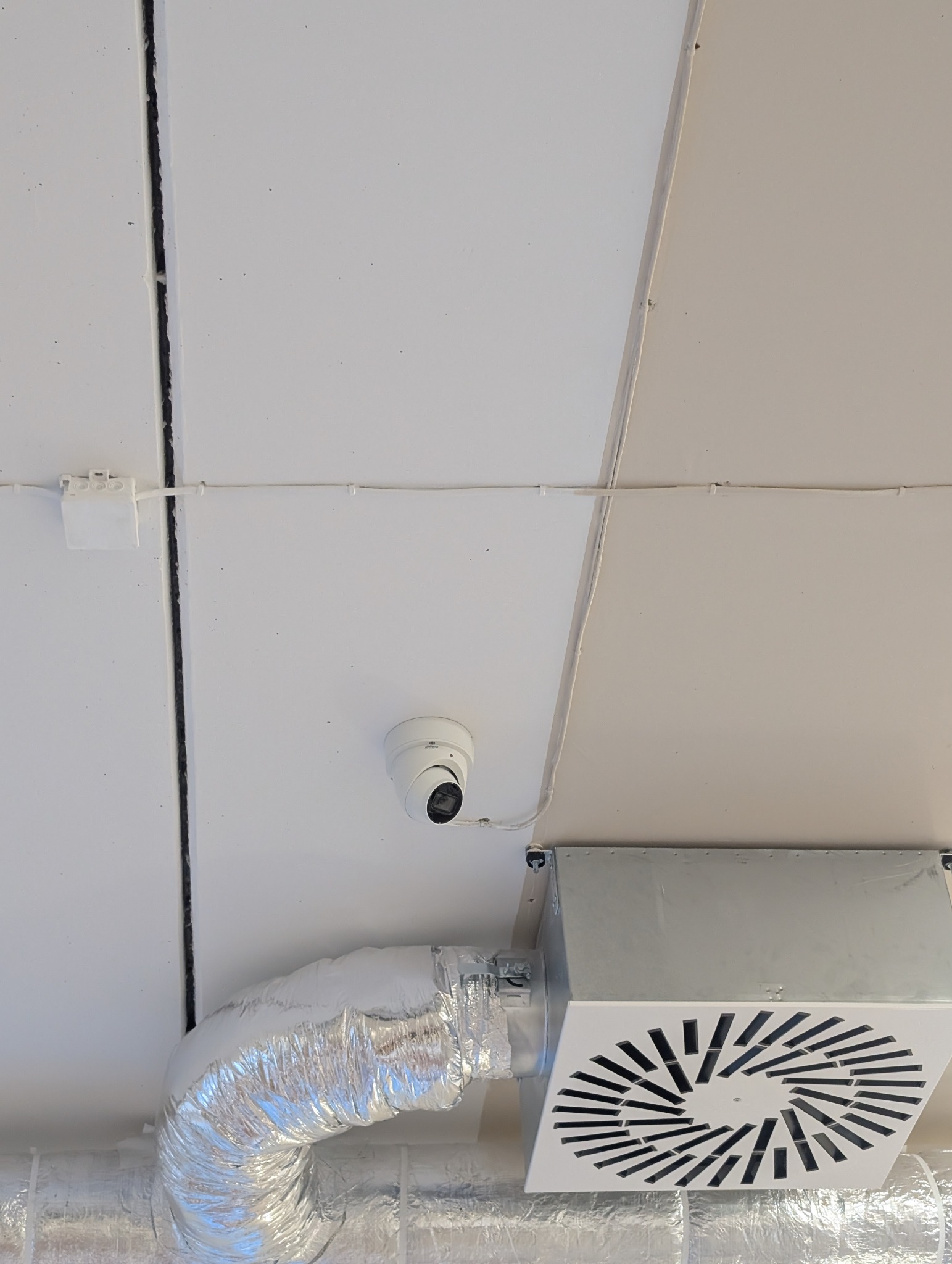 Biała kopułkowa kamera monitoringu zamontowana na suficie, obok metalowych kanałów wentylacyjnych. Widoczne przewody i puszka elektryczna.