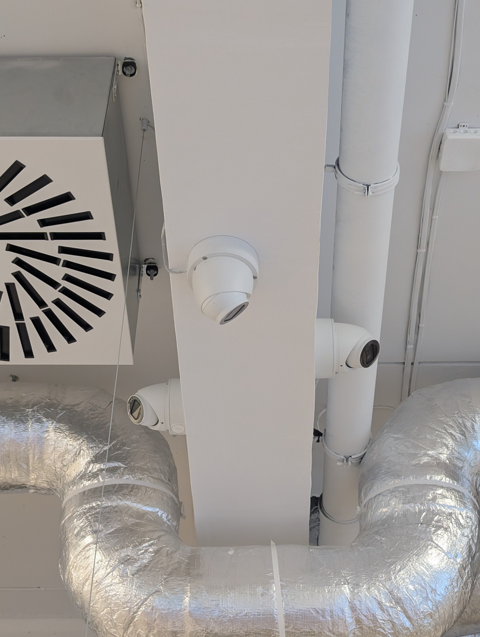 Sufit z zamontowanymi kamerami monitoringu kopułkowymi, widoczny system wentylacji i rury. Kamery skierowane w różnych kierunkach, instalacja na białym tle.