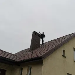 Mężczyzna w czarnym kombinezonie na dachu domu z brązową dachówką, czyści ceglany komin za pomocą długiego narzędzia, pochmurne niebo w tle.