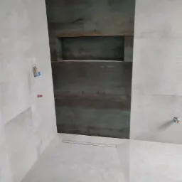 Surowa łazienka z szarymi płytkami imitującymi beton, akcentem ciemnobrązowej ściany z wnęką i odpływem liniowym w podłodze.