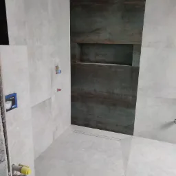 Wnętrze łazienki w trakcie remontu: szare płytki imitujące kamień, ciemna wnęka prysznicowa z półką, odpływ liniowy w podłodze, widoczne instalacje wodno-kanalizacyjne przed zabudową.