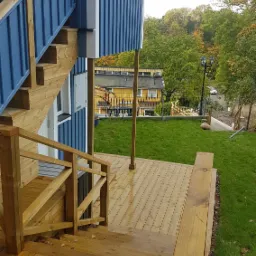 Drewniany taras z schodami przy niebieskim domu szkieletowym, widok na zielony trawnik i drzewa w tle.