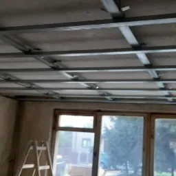 Montaż metalowej konstrukcji sufitu podwieszanego w remontowanym pokoju z oknem i drabiną