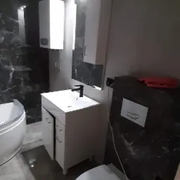 Wykończona łazienka z umywalką na szafce, wiszącą toaletą i wanną, płytki imitujące marmur na ścianach, widoczne niedociągnięcia w postaci niezamaskowanych przewod&oacute;w.