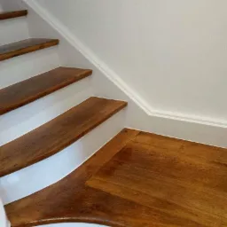 Drewniane schody z białymi podstopnicami i lakierowaną podłogą, wykończone białą listwą przypodłogową i dekoracyjną listwą na ścianie.