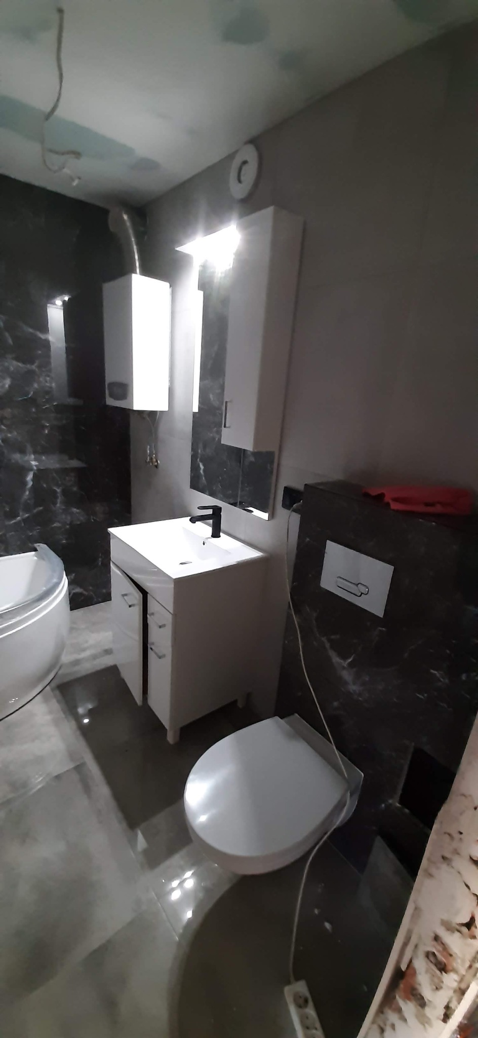 Wykończona łazienka z umywalką na szafce, wiszącą toaletą i wanną, płytki imitujące marmur na ścianach, widoczne niedociągnięcia w postaci niezamaskowanych przewodów.