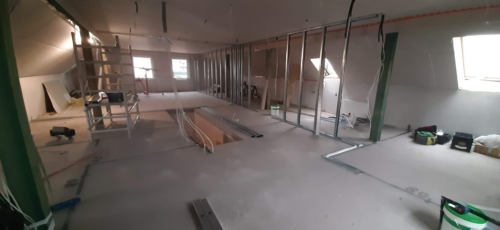 Poddasze w trakcie adaptacji: widoczne metalowe profile ścian działowych, otwór w podłodze z wystającymi przewodami, drabina, i narzędzia budowlane w nieładzie.