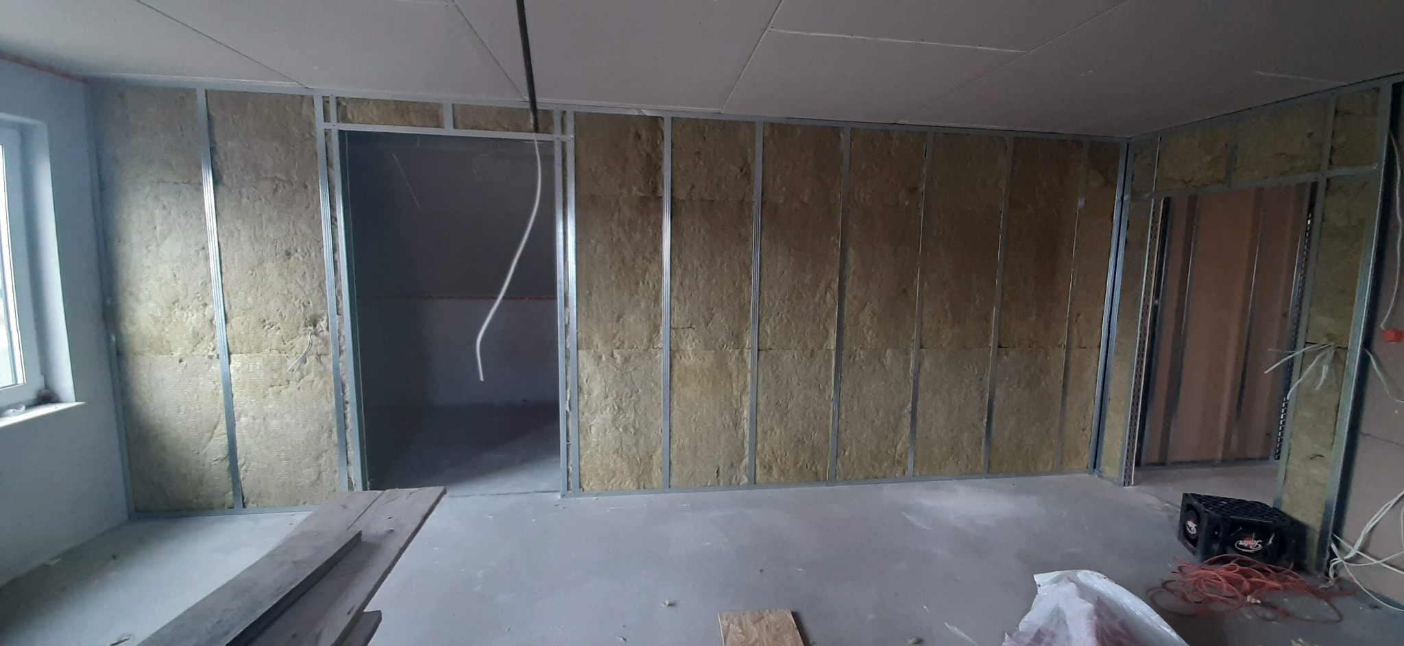 Wnętrze w trakcie prac: ściany z metalowym stelażem wypełnione wełną mineralną, widoczne przewody elektryczne, surowy stan wykończenia.
