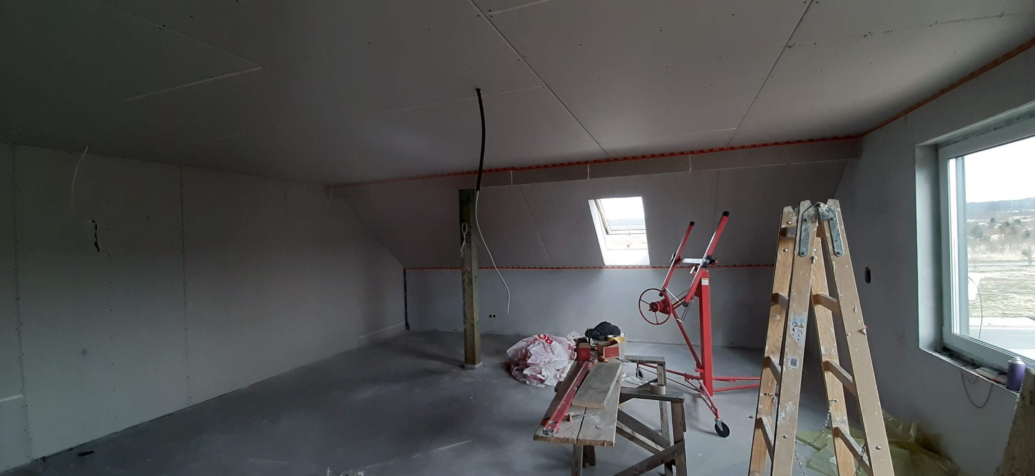 Poddasze w trakcie remontu: widoczne płyty gipsowo-kartonowe na ścianach i suficie, czerwona taśma miernicza, podnośnik do płyt, drabina i okno dachowe.