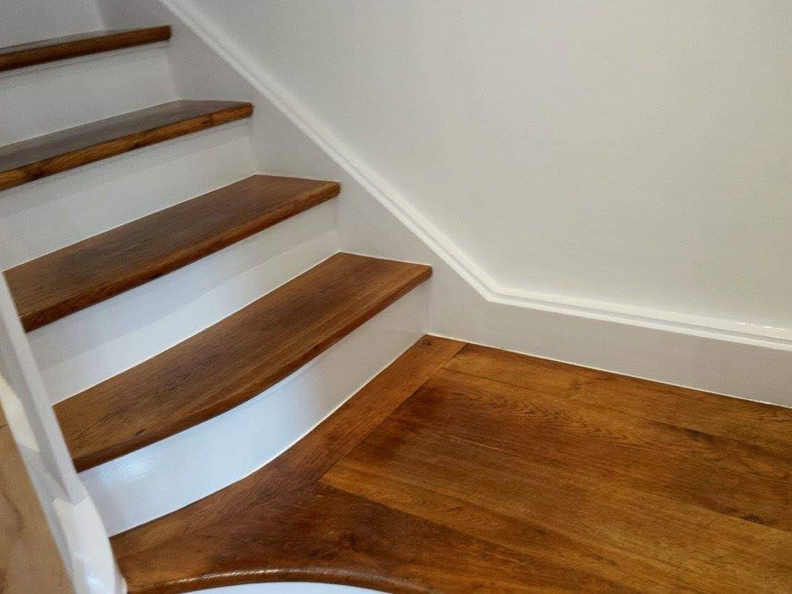 Drewniane schody z białymi podstopnicami i lakierowaną podłogą, wykończone białą listwą przypodłogową i dekoracyjną listwą na ścianie.