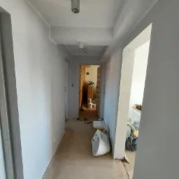 Korytarz po świeżym malowaniu na biało, widoczne ślady prac wykończeniowych, kartony i narzędzia, wnętrze w trakcie prac.