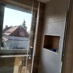 Nowoczesna kabina prysznicowa z widokiem na budynek mieszkalny w tle, płytki imitujące drewno na ścianach i wewnątrz wnęki, niebieska taśma malarska we wnęce.