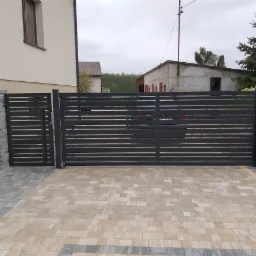Nowoczesna, przesuwna brama wjazdowa z poziomymi panelami, pomalowana na ciemny szary kolor, zamontowana na posesji z kostką brukową w odcieniach beżu i szarości. Widoczny fragment budynku z oknem...