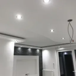 Nowoczesny sufit podwieszany z oświetleniem punktowym i elementami dekoracyjnymi na ścianie w odcieniach szarości i bieli. Widoczne niedokończone podłączenie lampy.