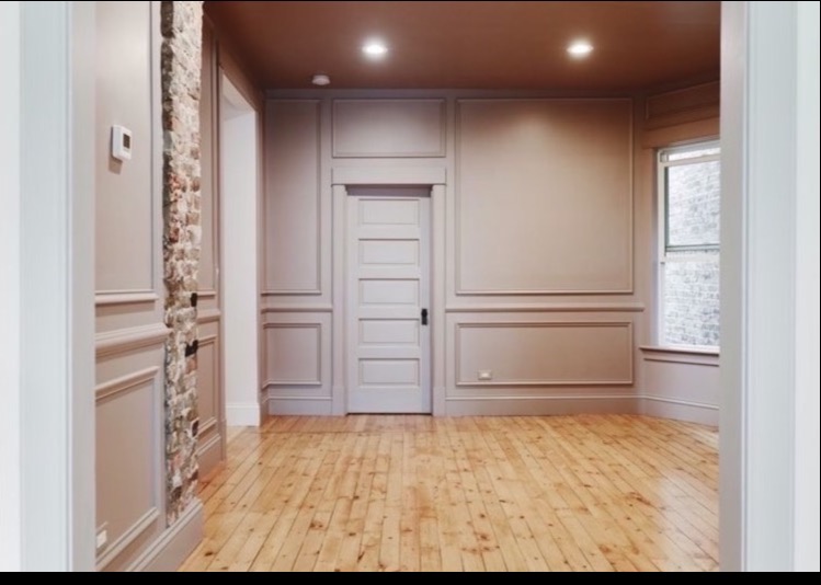 Pokój po remoncie z drewnianą podłogą, ścianami w odcieniach beżu z dekoracyjnymi panelami, odsłoniętym fragmentem ceglanej ściany i białymi drzwiami.