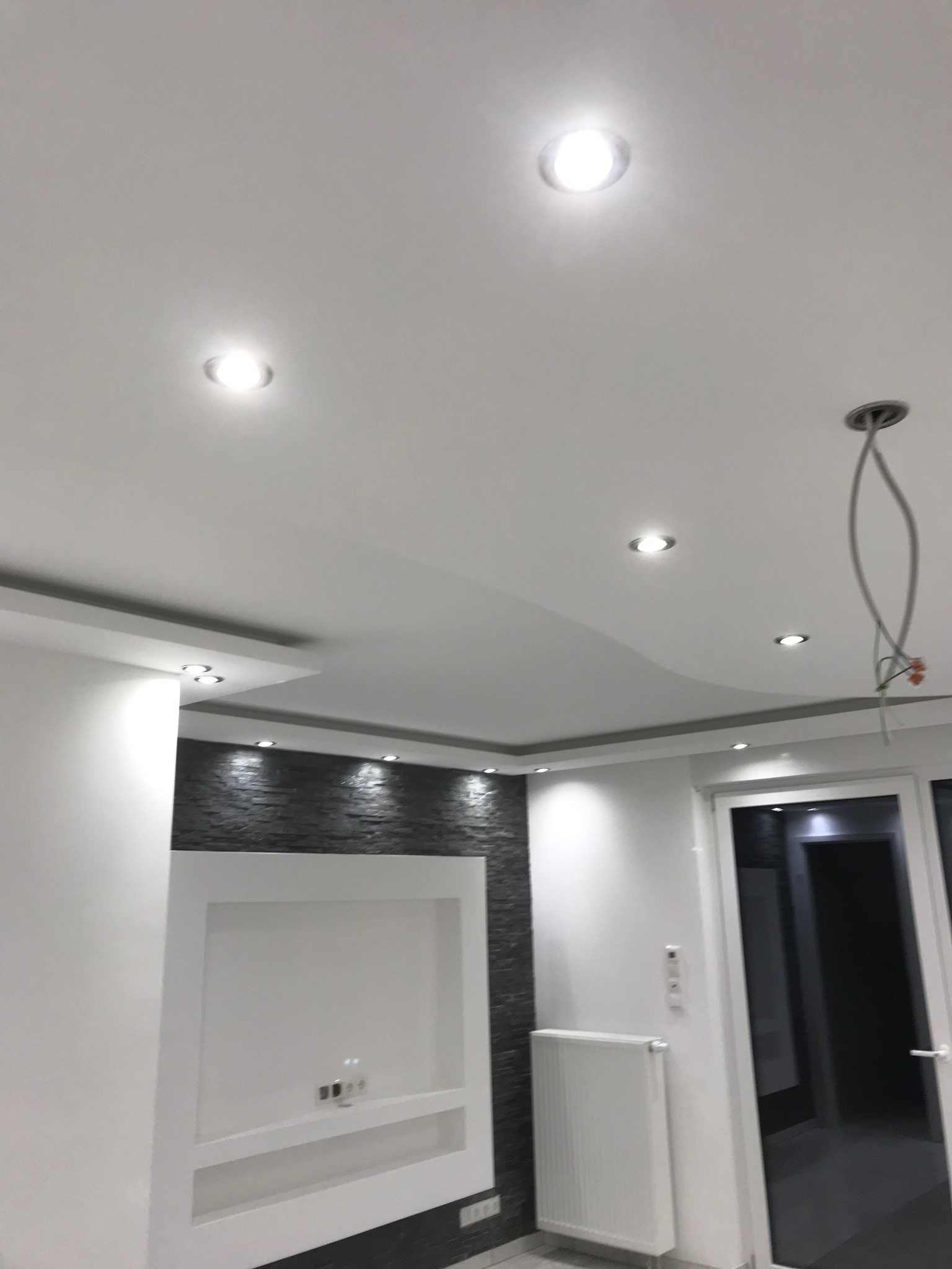 Nowoczesny sufit podwieszany z oświetleniem punktowym i elementami dekoracyjnymi na ścianie w odcieniach szarości i bieli. Widoczne niedokończone podłączenie lampy.