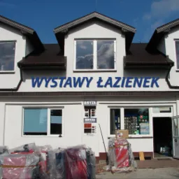 Fasada budynku z napisem 'Wystawy Łazienek' nad witrynami, przed wejściem zapakowane w folię piece i drabina oparta o ścianę.