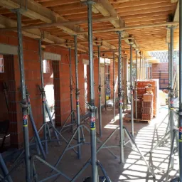 Wnętrze budowy domu z cegły ceramicznej, widoczne ściany w trakcie murowania, podparte metalowymi stemplami stropu, paleta z bloczkami ceramicznymi w tle.