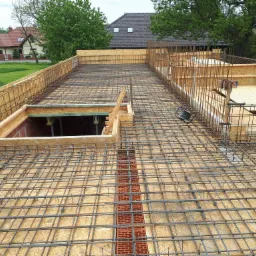 Zbrojenie stropu z siatki stalowej na szalunku z płyt OSB, widoczne otwory w stropie obudowane cegłą i drewnem, w tle domy jednorodzinne.