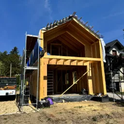 Konstrukcja szkieletowa domu w trakcie budowy, widoczne drewniane elementy, rusztowania i materiały izolacyjne na tle błękitnego nieba.