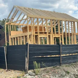 Wznoszenie drewnianej konstrukcji domu jednorodzinnego z widocznym szkieletem dachu i ścian, zabezpieczone czarną siatką na tle zieleni i błękitnego nieba.