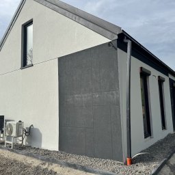 DOMY ENERGOOSZCZĘDNE, SZKIELETOWE TADEUSZ DORULA - Elewacja domu jednorodzinnego z fragmentem ściany wykończonej ciemnoszarą płytą elewacyjną. Widoczne klimatyzatory na zewnątrz budynku.