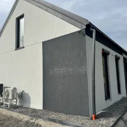 Elewacja domu jednorodzinnego z fragmentem ściany wykończonej ciemnoszarą płytą elewacyjną. Widoczne klimatyzatory na zewnątrz budynku.