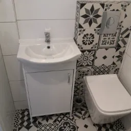 Wykończona łazienka w stylu retro z białą szafką pod umywalkę, wiszącą toaletą i dekoracyjnymi czarno-białymi płytkami podłogowymi i ściennymi, odbicie osoby robiącej zdjęcie w lustrze.