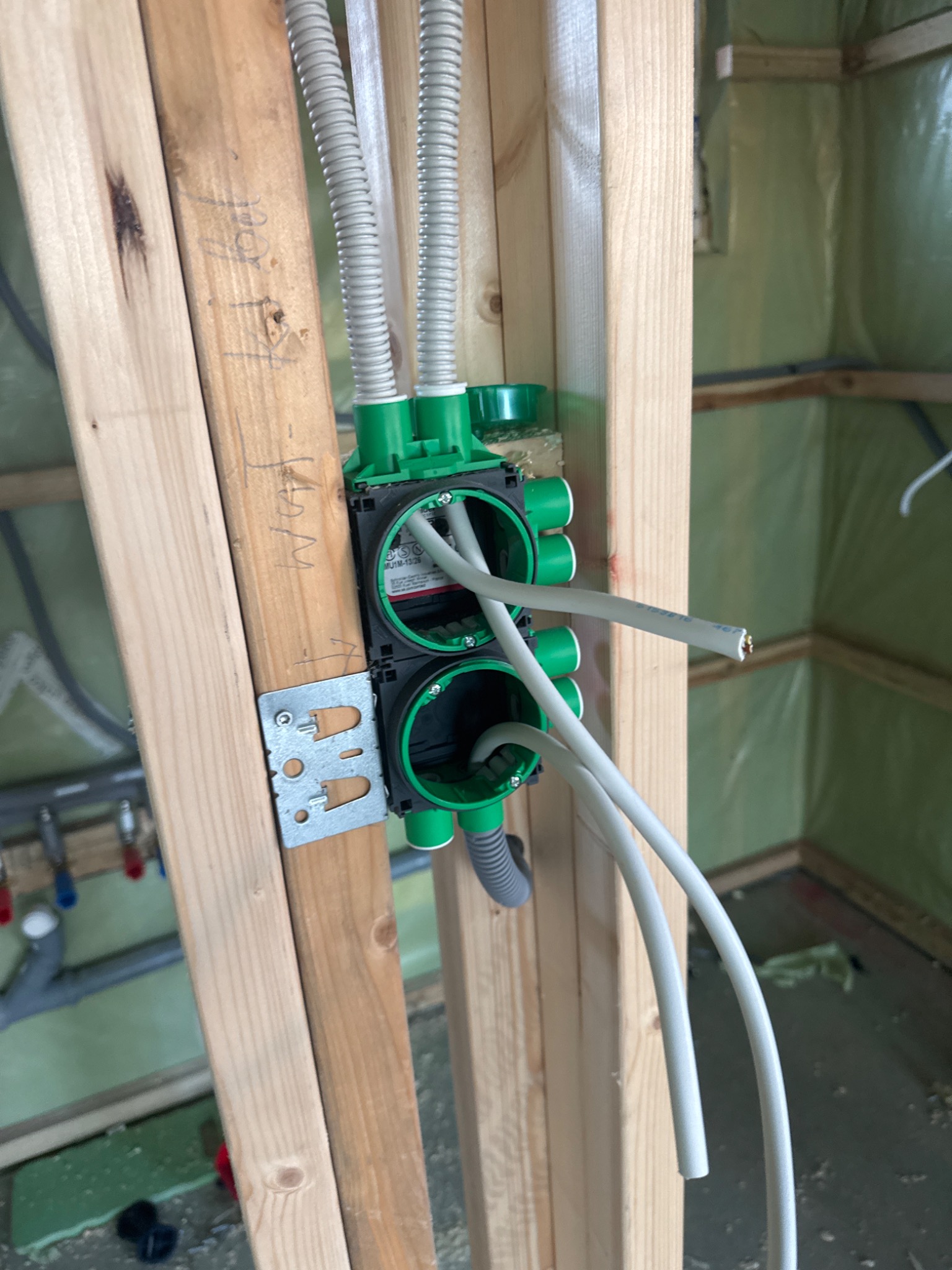 Zielona puszka instalacyjna z przewodami w drewnianej konstrukcji szkieletowej. Widoczne peszle i instalacja sanitarna w tle. Ściany zabezpieczone folią.