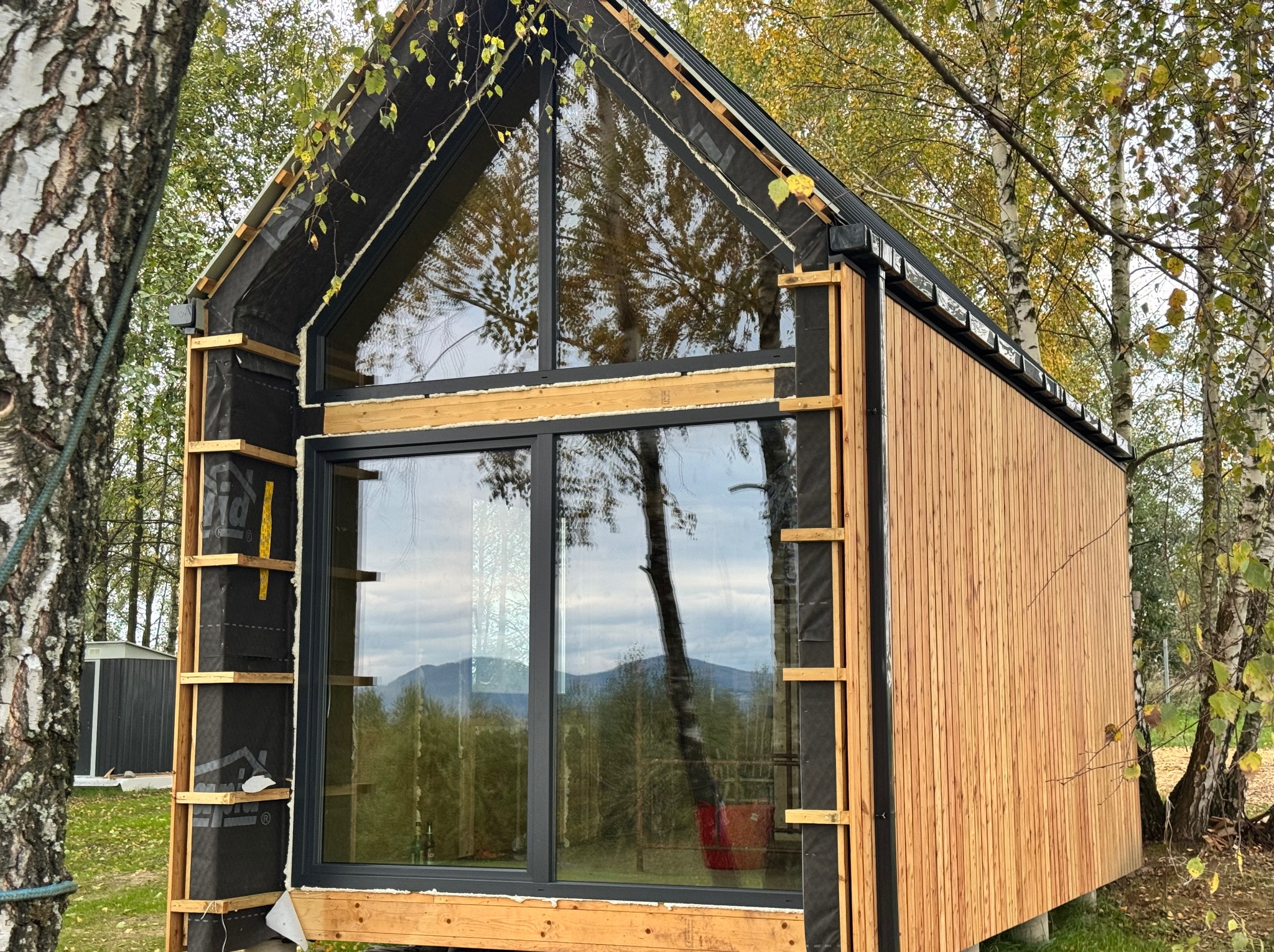 Dom z dużymi oknami w trakcie montażu elewacji drewnianej i izolacji. Widoczne rusztowania i elementy konstrukcyjne.