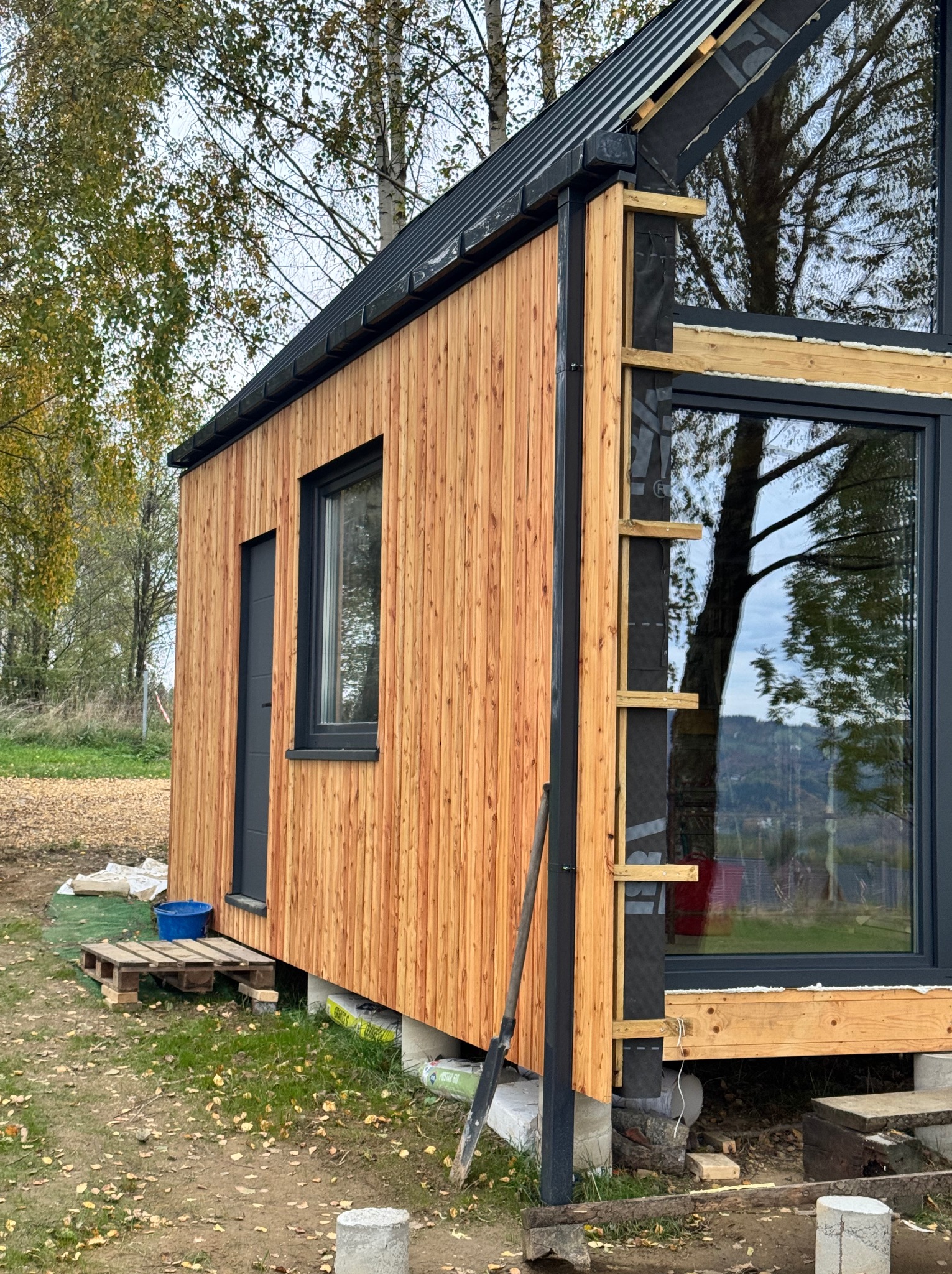 Nowoczesny dom z drewnianą elewacją w trakcie budowy. Widoczne okna, drzwi i elementy konstrukcyjne. Tło stanowią drzewa i trawa.