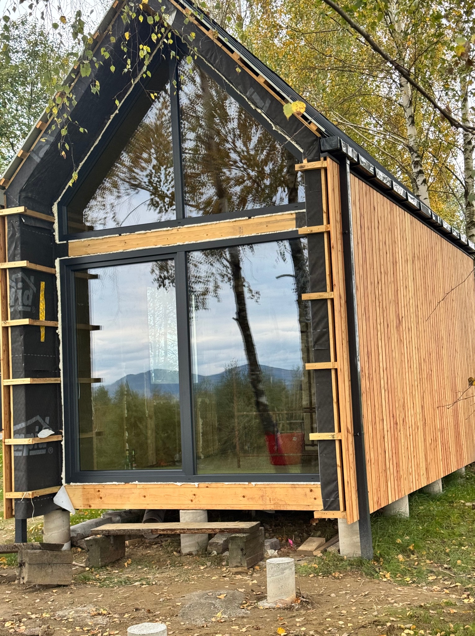 Nowoczesna elewacja domu w trakcie budowy: drewniana okładzina, duże okna z widokiem na góry. Widoczne detale konstrukcyjne i izolacja. Jesienne liście na ziemi.