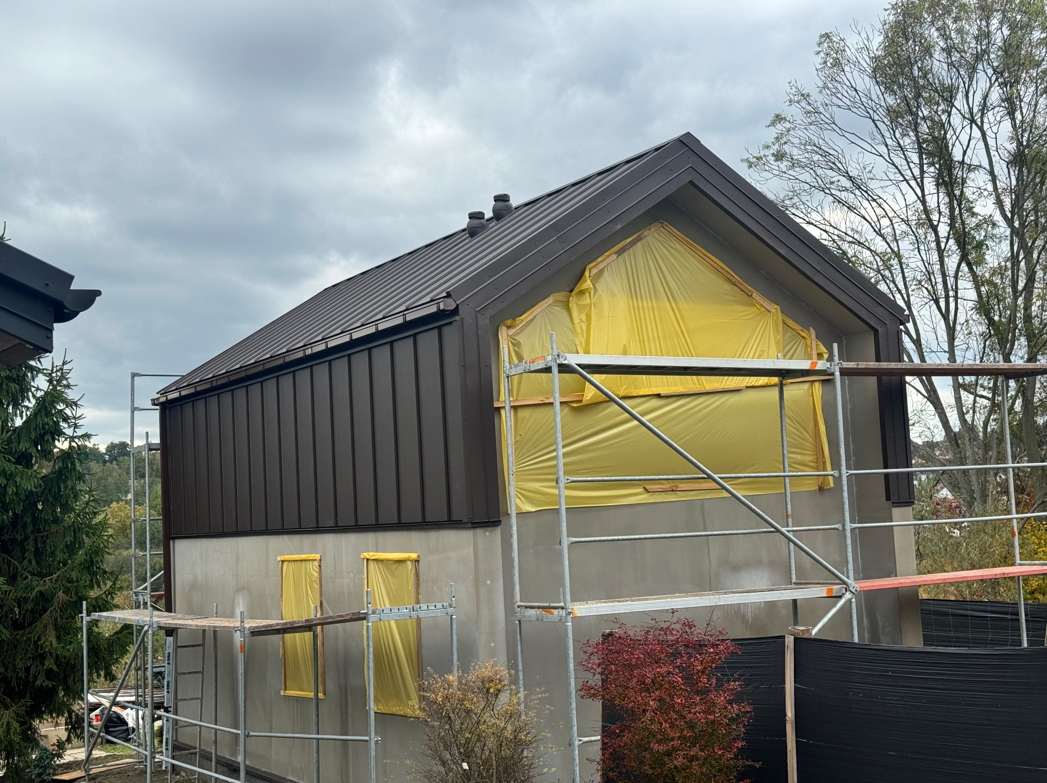 Dom w trakcie prac wykończeniowych: szare ściany, brązowy dach z blachy na rąbek stojący, okna zabezpieczone folią, rusztowania. Widok z dołu na budynek.