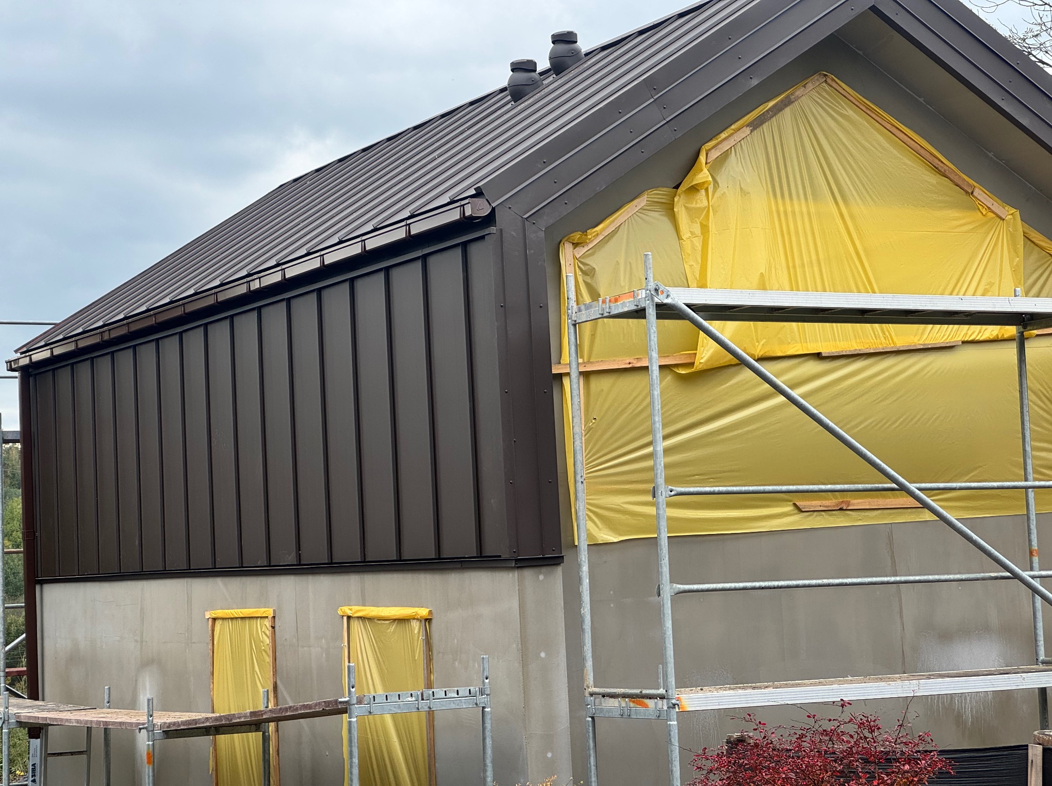 Dom w trakcie prac wykończeniowych: brązowe panele elewacyjne, metalowy dach, rusztowania i folia ochronna na oknach. Widok na fragment budynku w pochmurny dzień.