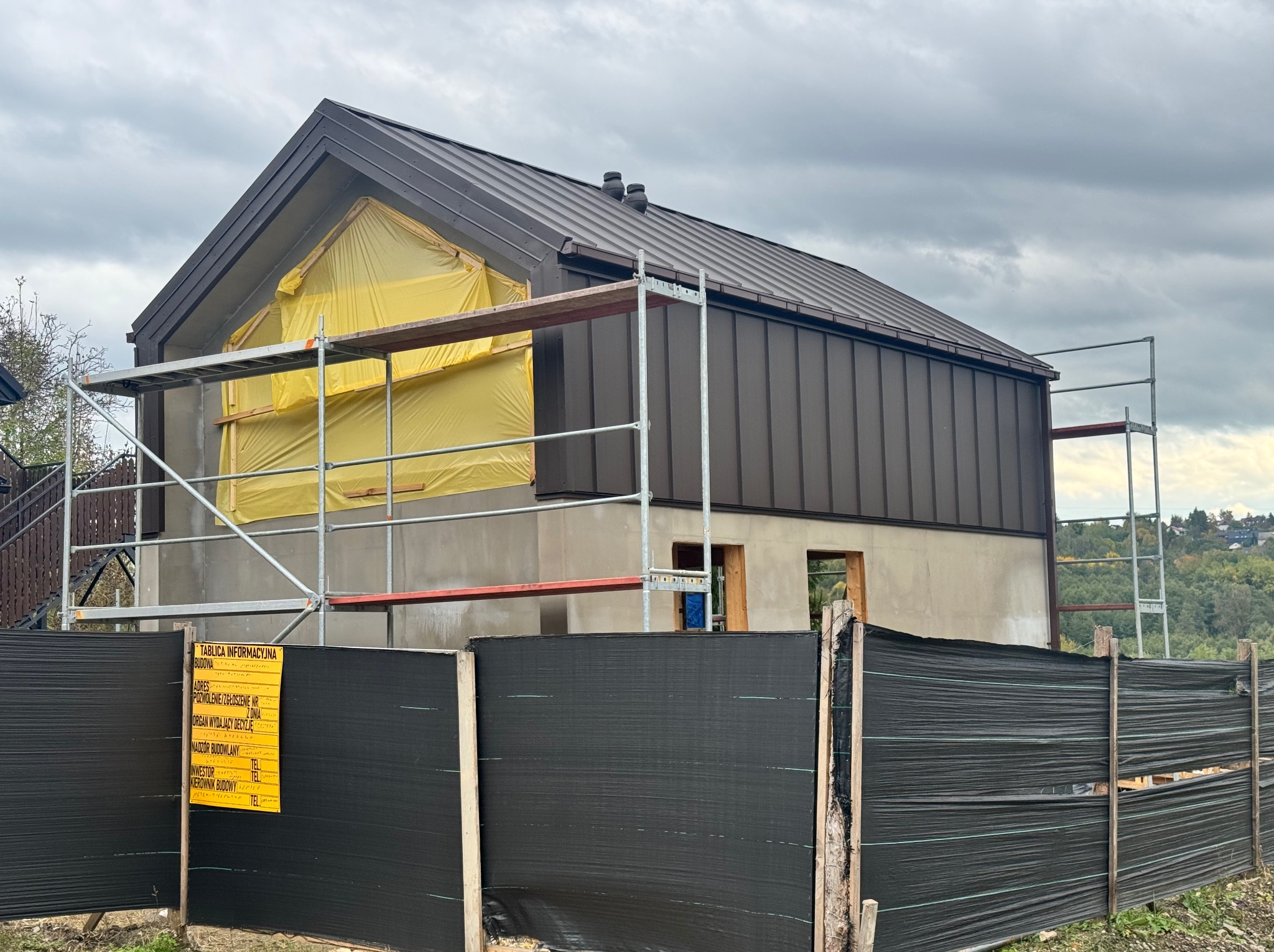 Dom w trakcie ocieplania: elewacja z rusztowaniem i folią ochronną na oknach, brązowy dach z blachy na tle pochmurnego nieba. Widoczna tablica informacyjna budowy.
