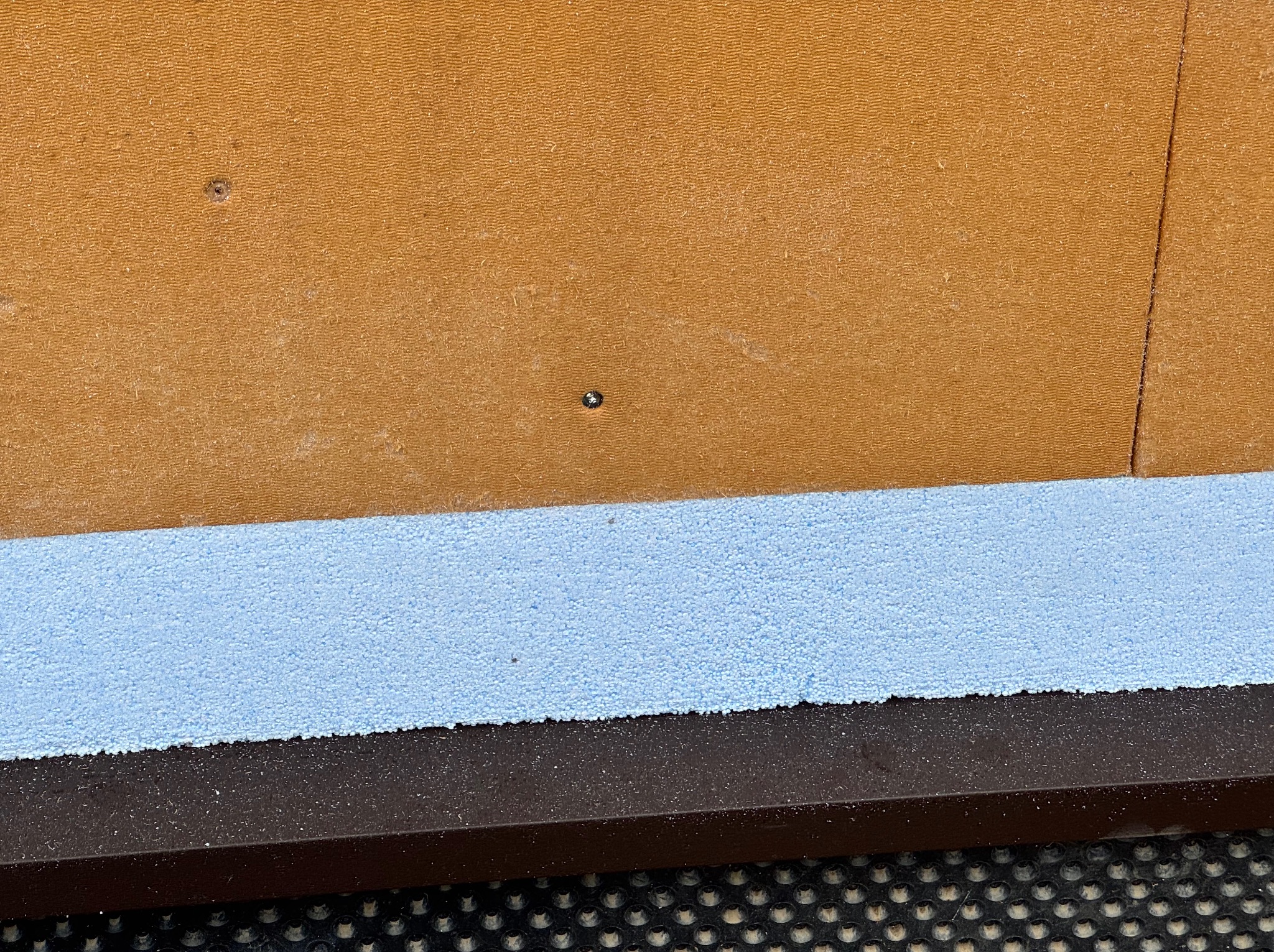 Fragment ściany z płytą OSB, izolacją z jasnoniebieskiego styropianu i brązową listwą przypodłogową na czarnej macie z okrągłymi otworami.