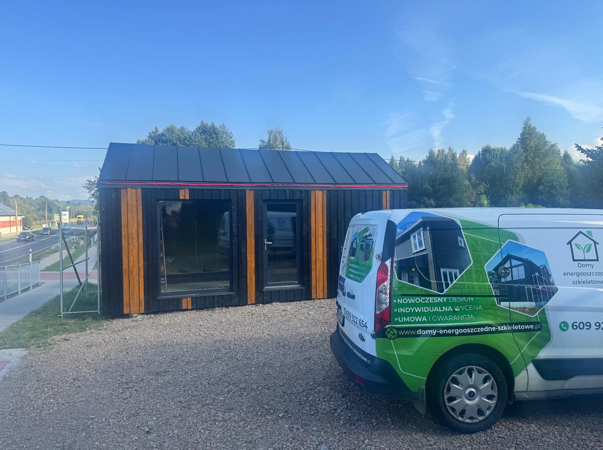Gotowy, energooszczędny dom szkieletowy z czarnym dachem i drewnianą fasadą, obok firmowy bus z reklamą. Nowoczesny design i indywidualna wycena.