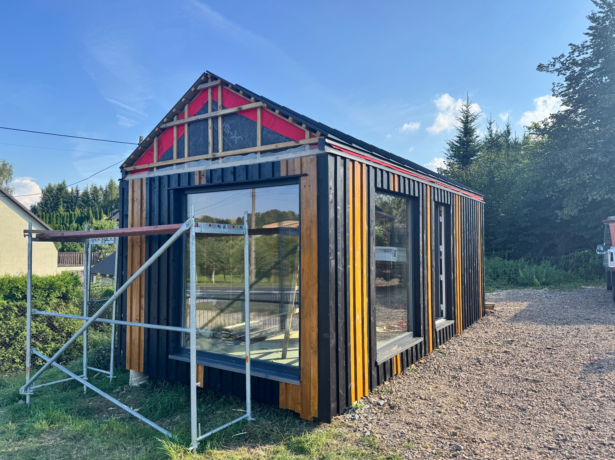 Nowoczesny budynek w trakcie budowy z czarno-żółtą elewacją, widoczne rusztowania i elementy konstrukcyjne dachu. Słoneczny dzień.